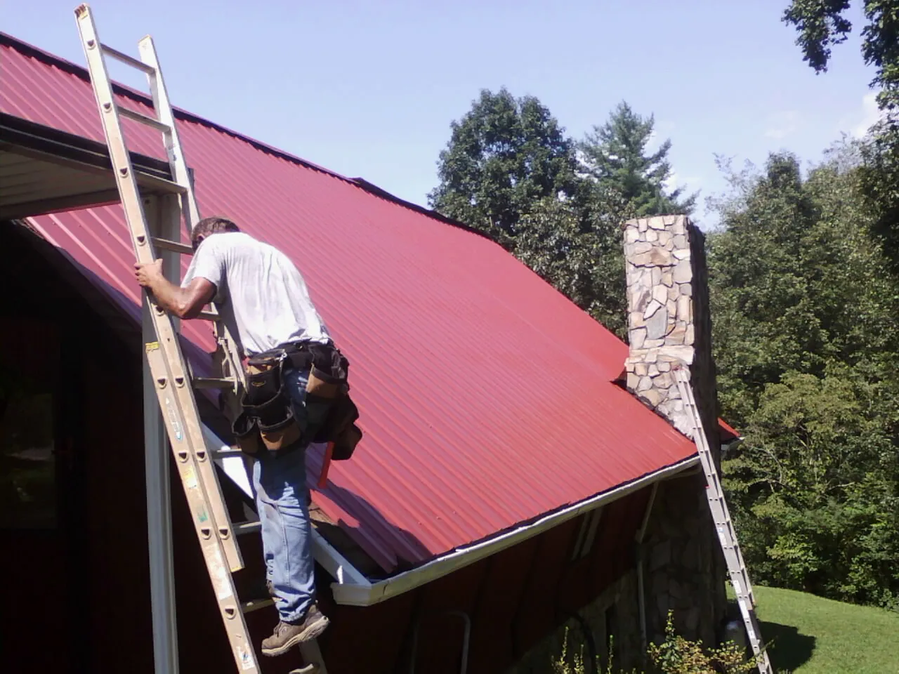 Roofer on a ladder beside a red metal roof and stone chimney — Soffit & Fascia Repair by Roofing Brothers in Bethel Springs
